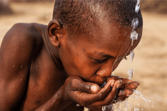 Ein Junge trinkt aus den Händen frisches Wasser aus einem Wasserlauf, symbolisiert den Zugang zu lebenswichtigen Ressourcen.
